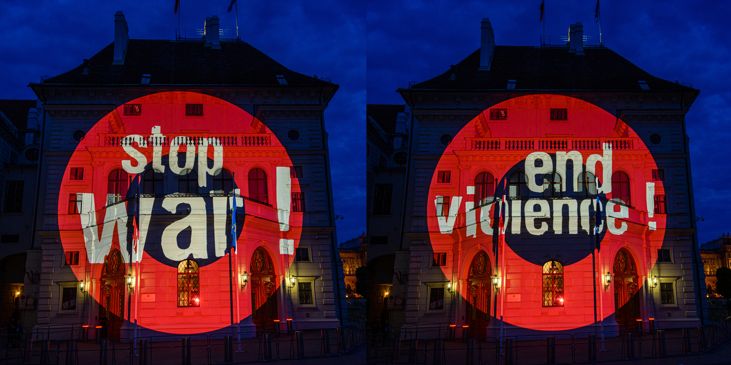 Eine nächtliche Wandprojektionen, in je einem roten Kreis sieht man die Sätze "Stop war!" und "End violence!".