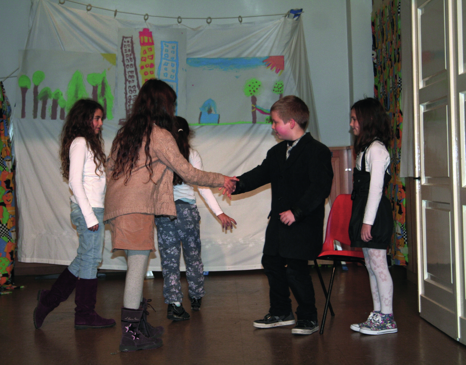 5 Theater spielende Kinder, ein Mädchen gibt einem Buben in schwarzem Anzug die Hand, die anderen Kinder tragen Alltagskleidung, im Hintergrund ein gespanntes weißes Leintuch, auf dem ein Wald, eine Wiese und in der Mitte Hochhäuser gemalt it