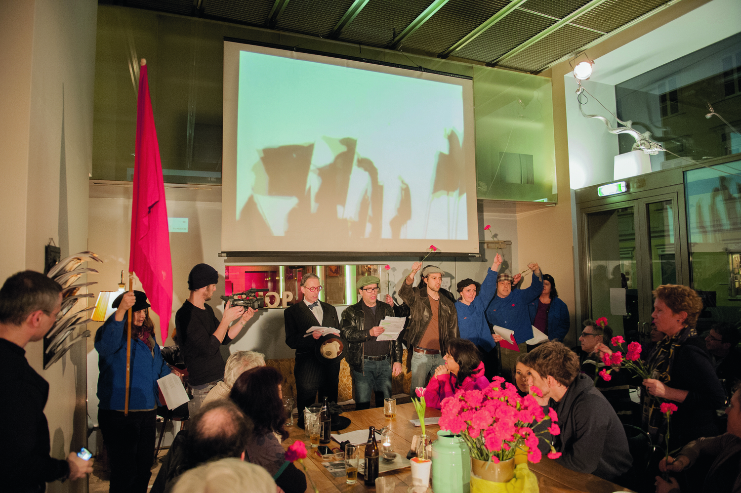 Menschen stehen an einem Tisch, an dem auch Menschen sitzen, und singen, dahinter erkennt man eine Projektion auf einer Stehleiwand