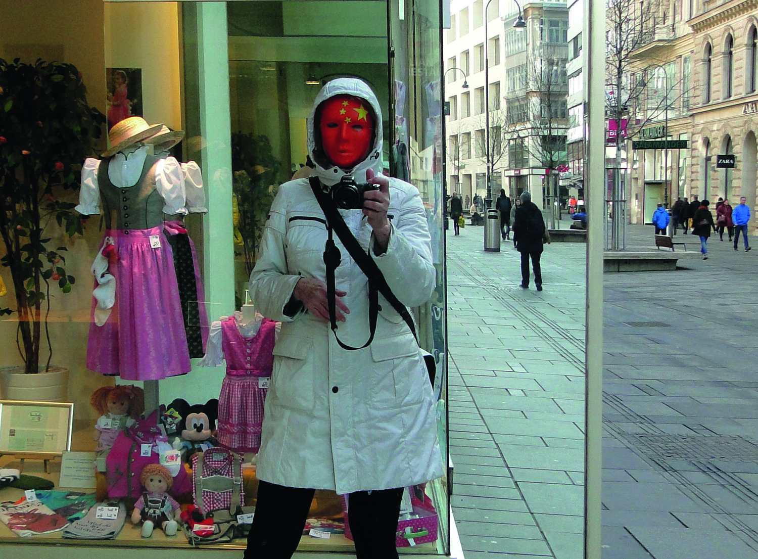 Eine Person mit roter Gesichtsmaske und im weißen Wintermantel fotografiert sich selbst in einen Spiegel eines Wiener Geschäftslokals (Kärntnerstraße) blickend