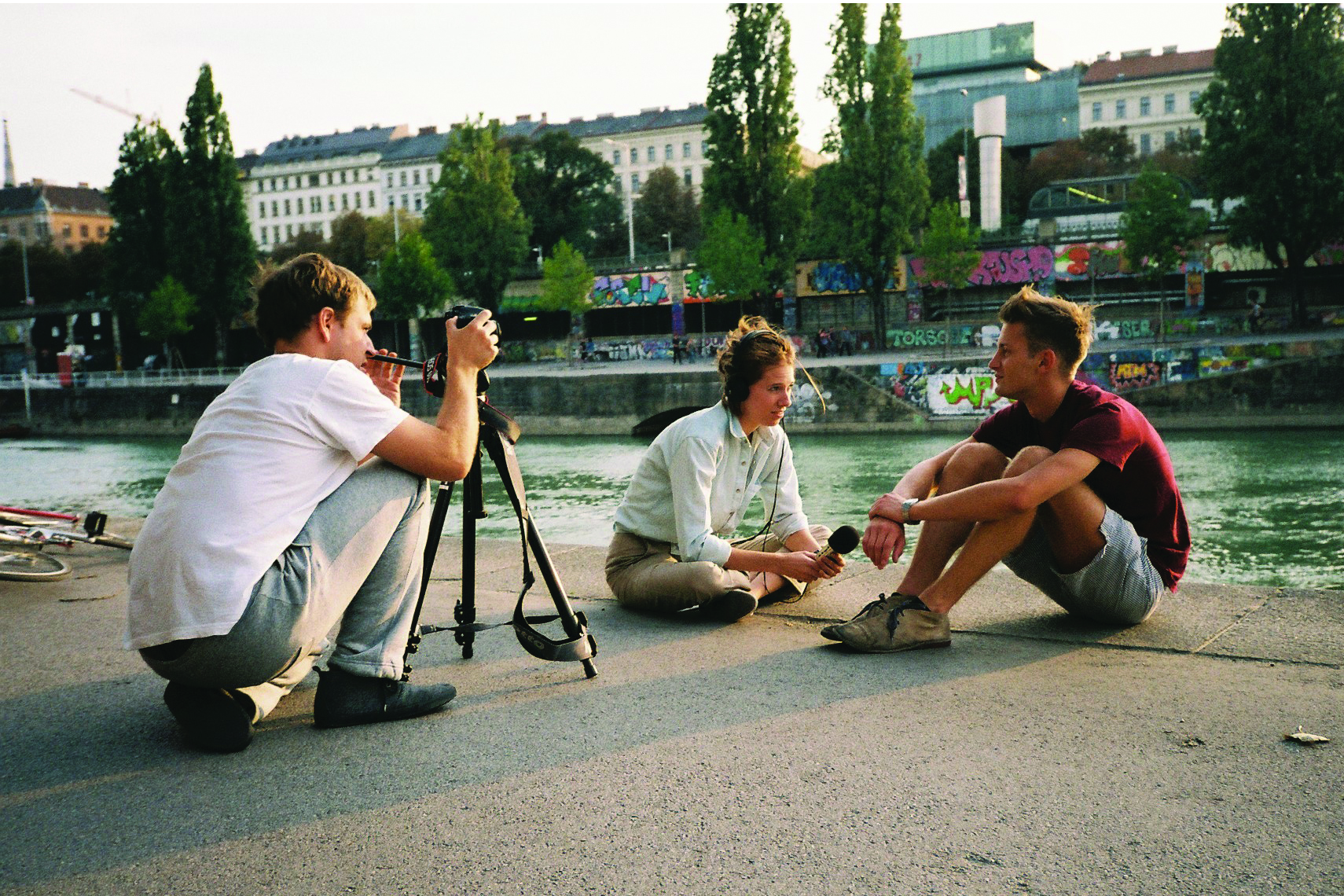 3 Personen sitzen am Ufer des Wien-Flusses und plaudern, einer fotografiert dabei