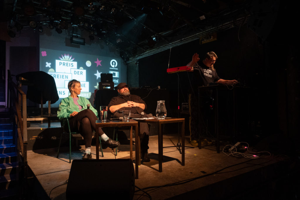 Susi Rogenhofer und Günther Friesinger auf der Bühne um einen Tisch sitzend bei der Moderation der Veranstaltung zum Preis der freien Szene Wiens 2024, daneben Jopa Jotakin am Theremin.