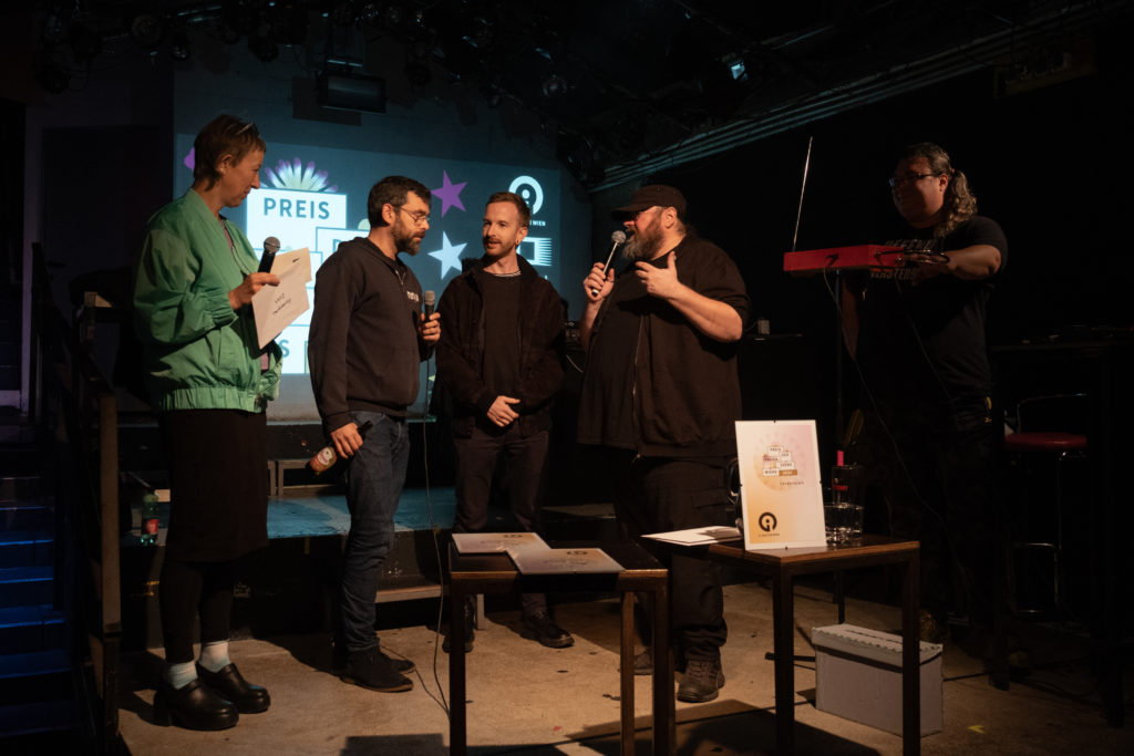 Auf der Bühne von links nach rechts: Susi Rogenhofer, zwei Vertreter*innen des Gewinners der Förderpreises Venster9, Günther Friesinger (in Mikrofon sprechend) und im Schatten Jopa Jotakin hinter seinem Theremin-