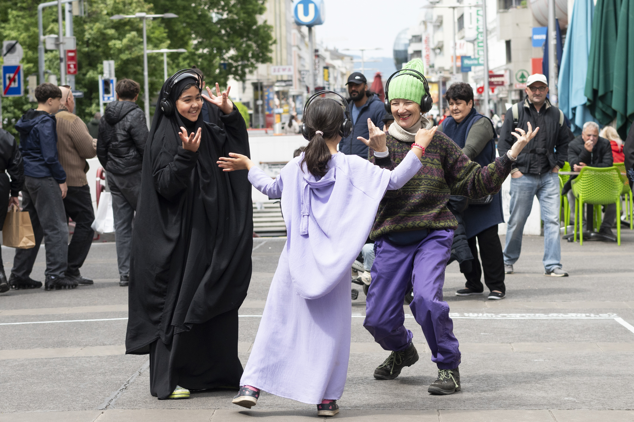 "Kinder und eine Performerin tanzen miteinender. sie tragen Kopfhörer und lachen beim Tanzen. Dahinter sind Passant:innen und schauen zu oder gehen grade vorbei. das ältere mädchen trägt eine Hidschab schwarz, das jüngere ein hell violettes langes Kleid. Die Performerin eine grüne Zwergenhaube und eine violette Hose