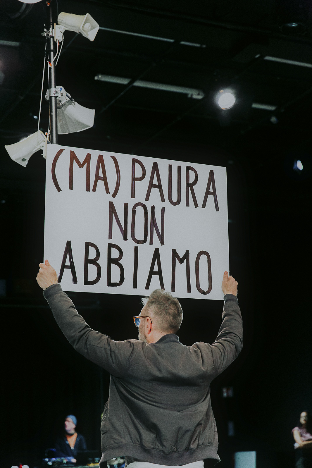Ein Performer (Aldo Giannotti) hält eines der vielen im Stück verwendeten Protestschilder mit der Message 'wir haben keine Angst' - eine Einladung an das Publikum am Ende des Stückes, gemeinsam einen Song zu singen.