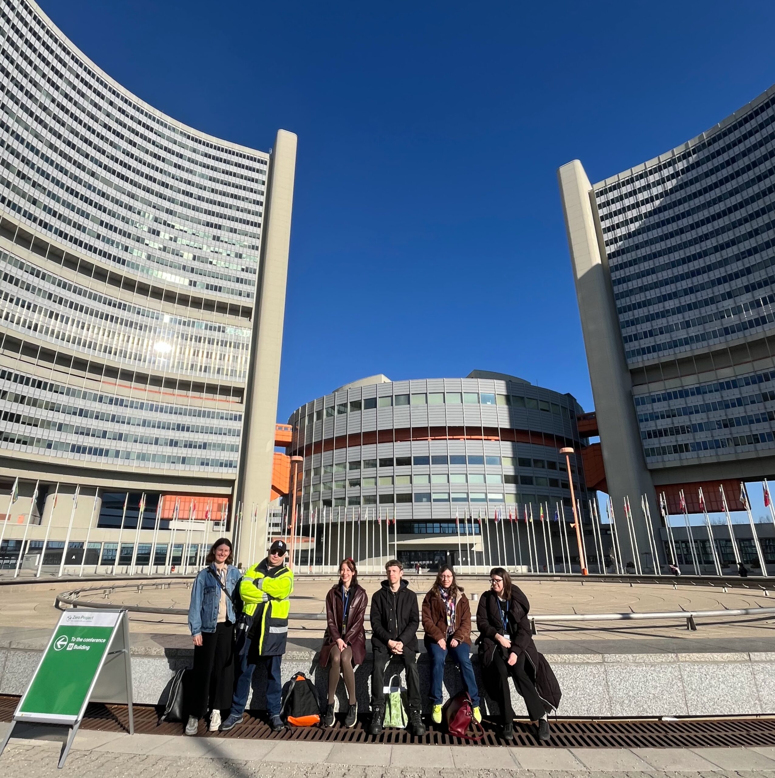 Vor den großen Gebäuden der UNO-City in Wien sitzen und stehen mehrere Personen. Der Himmel ist blau, die Personen sind teils geblendet durch die Sonne und lächeln.
