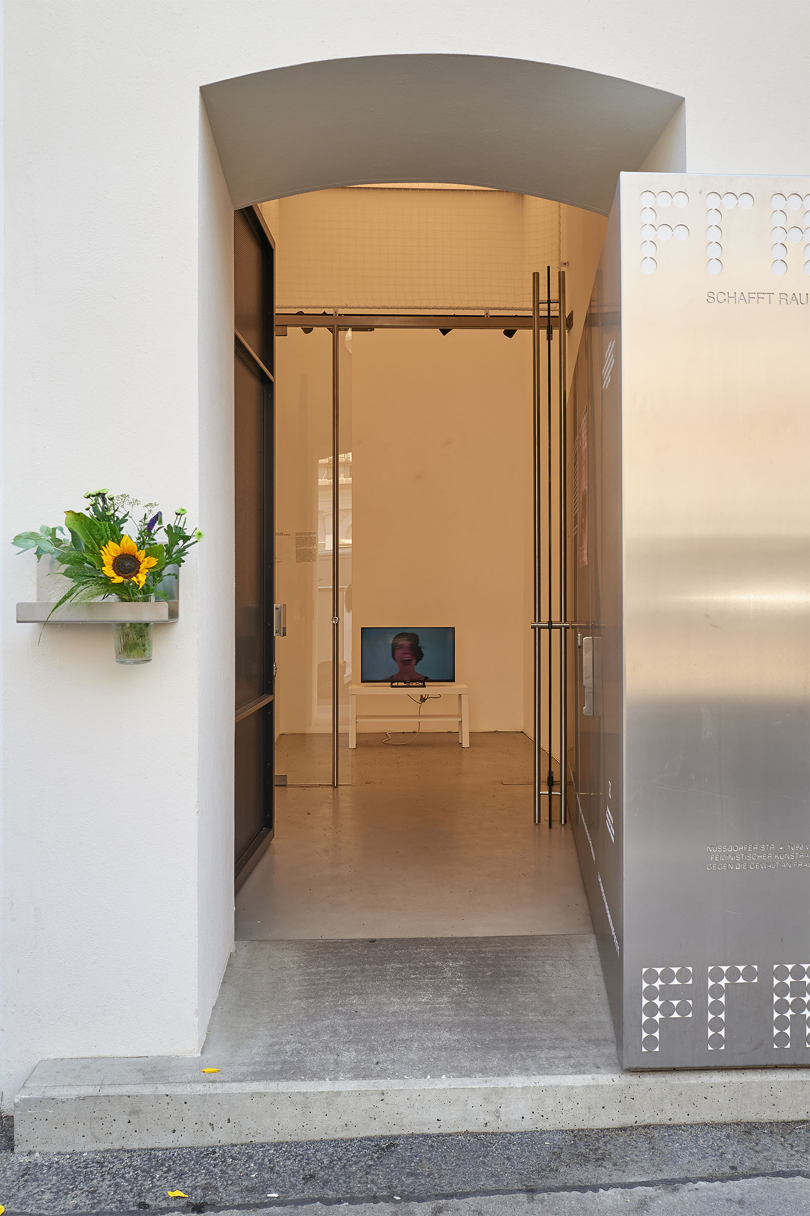 Die Fotografie zeigt den Blick von außen in den Ausstellungsraum FRAU SCHAFFT RAUM. Durch den offenen Eingang ist im Inneren ein Bildschirm auf einem weißen Tisch zu sehen, auf dem eine Videoarbeit mit einer Person läuft. Links neben dem Eingang steht ein kleiner Strauß mit Sonnenblume, rechts eine Metalltafel mit dem Schriftzug des Ausstellungsorts.