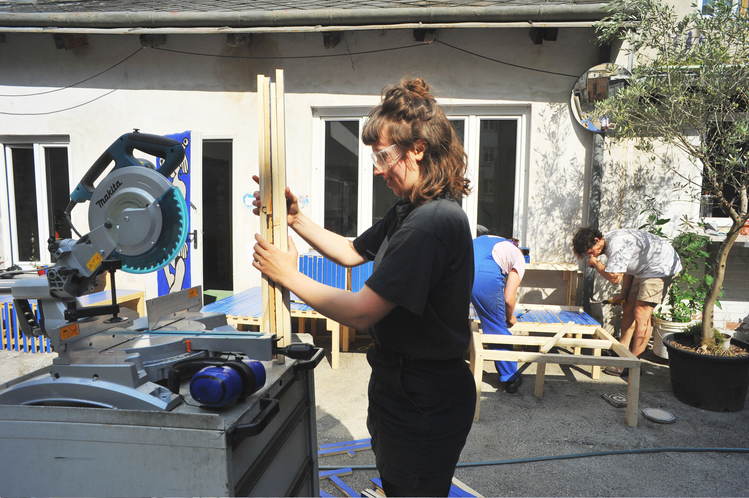 Das Bild zeigt einen sonnigen Innenhof, in dem drei Personen gemeinsam an einem Holzbau-Projekt arbeiten. Im Vordergrund hält eine Person mit Schutzbrille mehrere Holzlatten in der Hand, vor einer elektrischen Säge. Im Hintergrund bauen zwei weitere Personen an einem Rahmen aus Holzleisten – eine davon trägt einen blauen Overall. Die Szene wirkt handwerklich, gemeinschaftlich und kreativ. Der Ort hat eine helle Fassade mit großen Fenstern, ein bemaltes Türpaneel und einige Pflanzen in Töpfen. Die Stimmung ist konzentriert und kooperativ.