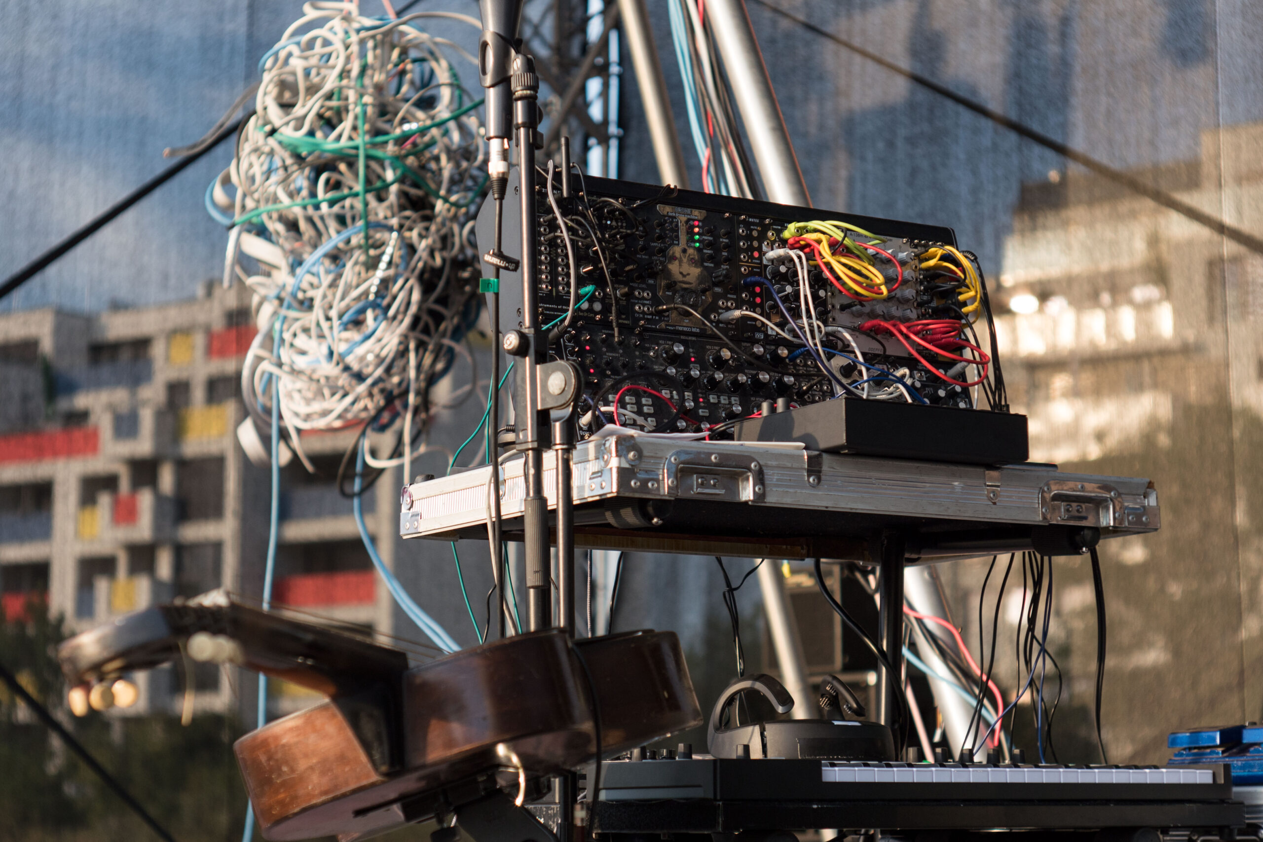 Der Apparatus (hier in Form eines Kabelkläuls mit ca. 1m Durchmesser) hängt von der Decke in der Luft. Vor ihm rechts im Bild stehen der modulare Synthesizer, weitere elektronische Instrumente, das Brain Computer Interface und eine alte Wiener Stahlsaitengitarre aus 1950, welche um einen digitalen Controller erweitert wurde und horizontal auf einem Ständer montiert gespielt wird.