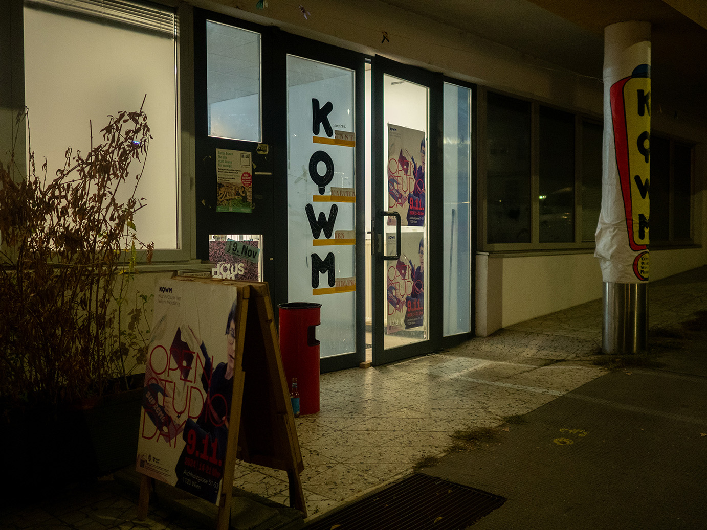 Eingang des KunstQuartier Wien Meidling. Es ist Nacht. Licht dringt durch die Glastüren und Fenster, die mit Plakaten und den Buchstaben K, Q, W und M beklebt sind. Vorne links steht ein Plakatständer mit einem Plakat, das auch an der Tür zu sehen ist. Weiter links davon ist eine Pflanze zu sehen. Rechts im Bild eine Säule auf der ebenfalls die Buchstaben K, Q, W und M zu lesen sind.