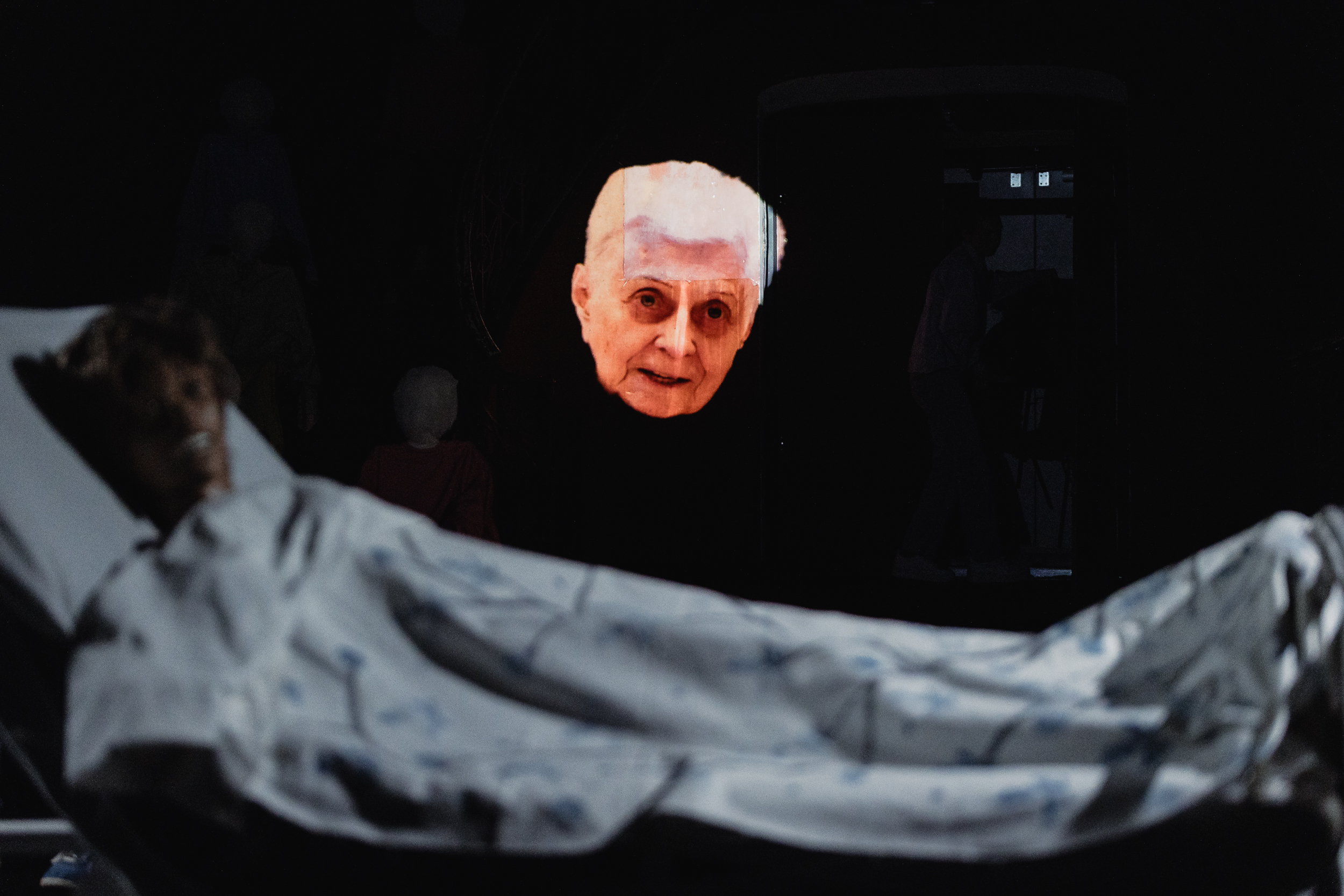 Das Bild zeigt eine Bewohnerin, die zum Publikum spricht. Im Vordergrund liegt eine Pflegepuppe, an der pflegerische Handgriffe erlernt werden. Zwei Realitäten treffen aufeinander.
