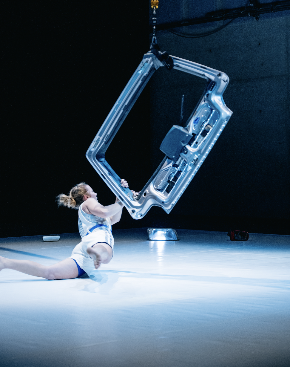 Die Performerin liegt halb auf dem weißen Tanzboden, halb hält sie sich an der Heckklappe fest. Dieses ist mir einem Seil nach oben gehängt. Im Hintergrund sehen wir eine ausgebaute Autoleuchte. Die Performerin ist von uns abgewendet und blick zum heck.