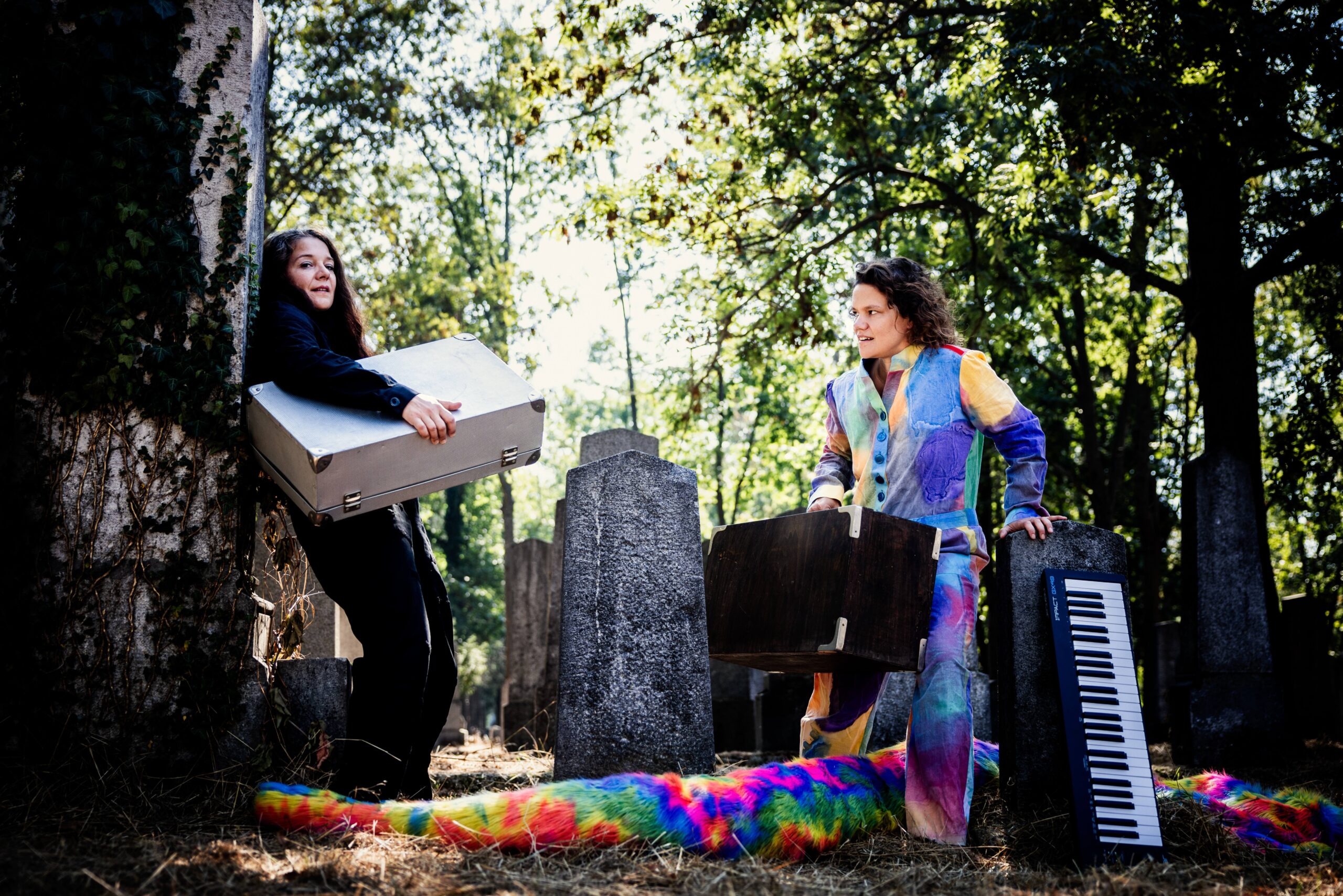 Regina Picker lehnt links im Bild mit einem silbernen Koffer in der Hand an einem Grabstein, während Julia Schreitl recht im Bild mit einem braunen Holzkoffer über das regenbogenfarbige Kuschelkostüm steigt. Neben ihr lehnt auch ein Keyboard an einem Grabstein.