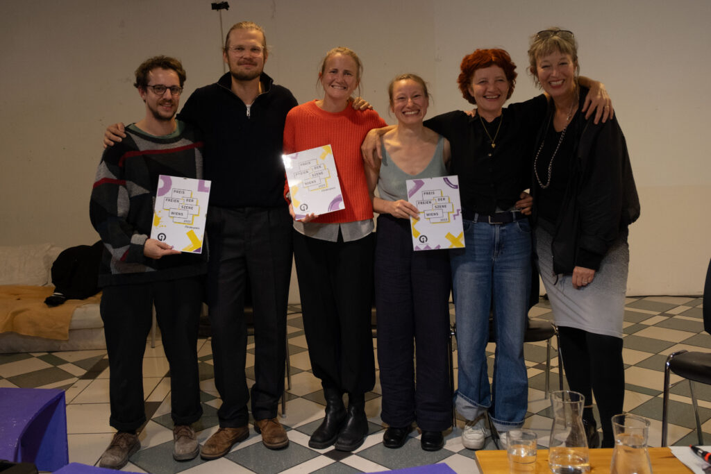 Die Preisträger*innen (5 Personen) und die Moderatorin stehen nebeneinander uns zeigen die Preisurkunden in die Kamera.